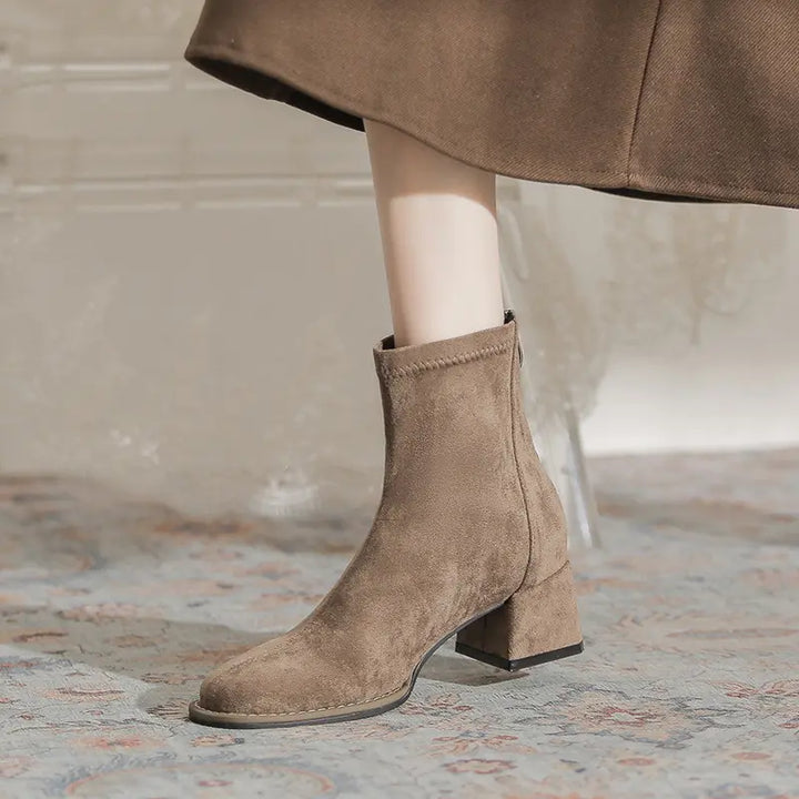 A pair of chic, taupe suede-like ankle boots with a square toe and block heel, shown on a leg wearing a brown skirt, styled for autumn/winter.