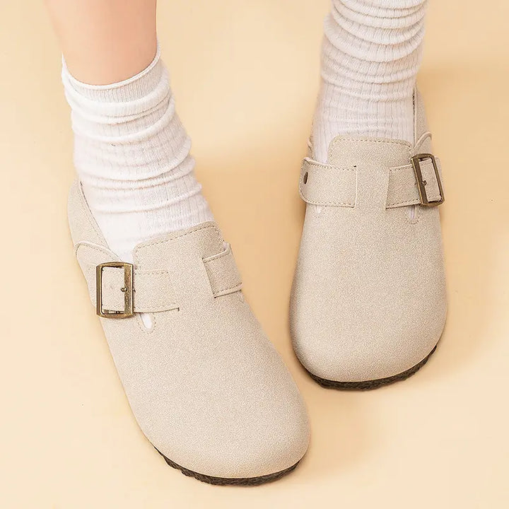 A pair of taupe suede-effect slip-on mules with adjustable antique brass buckles, worn on feet with white crew socks. These retro casual shoes feature a round toe and flat sole, presented against a soft yellow backdrop.