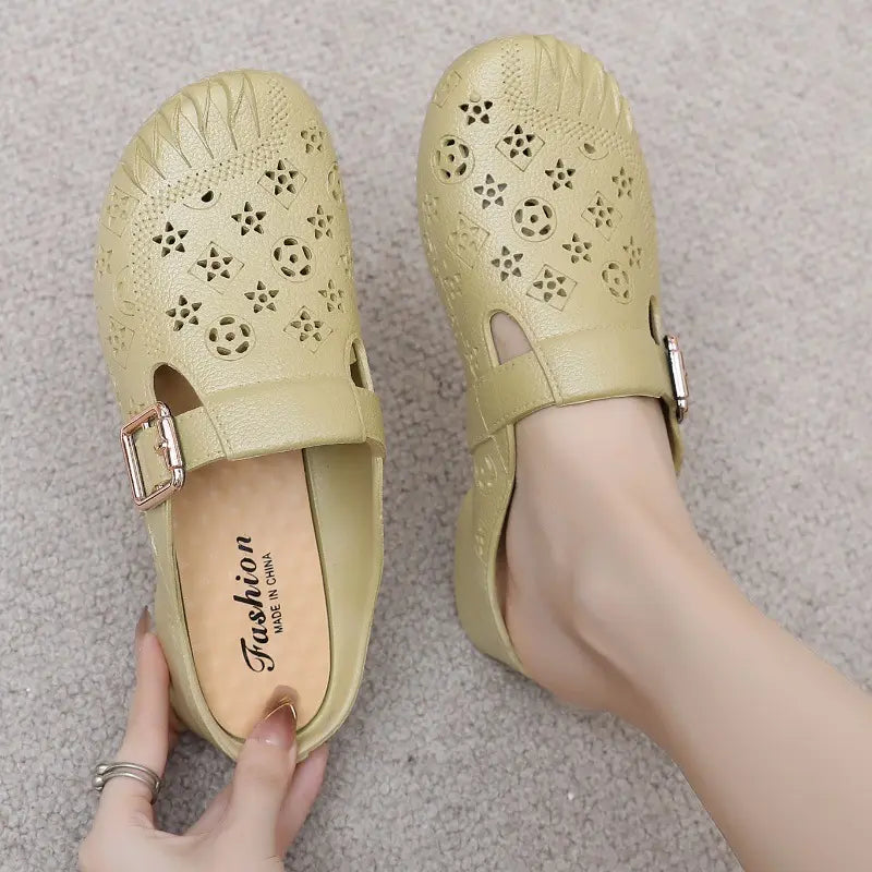 A pair of textured synthetic mules in a light beige color, featuring an embossed pattern, cut-out details, and an adjustable gold-tone buckle strap across the vamp.