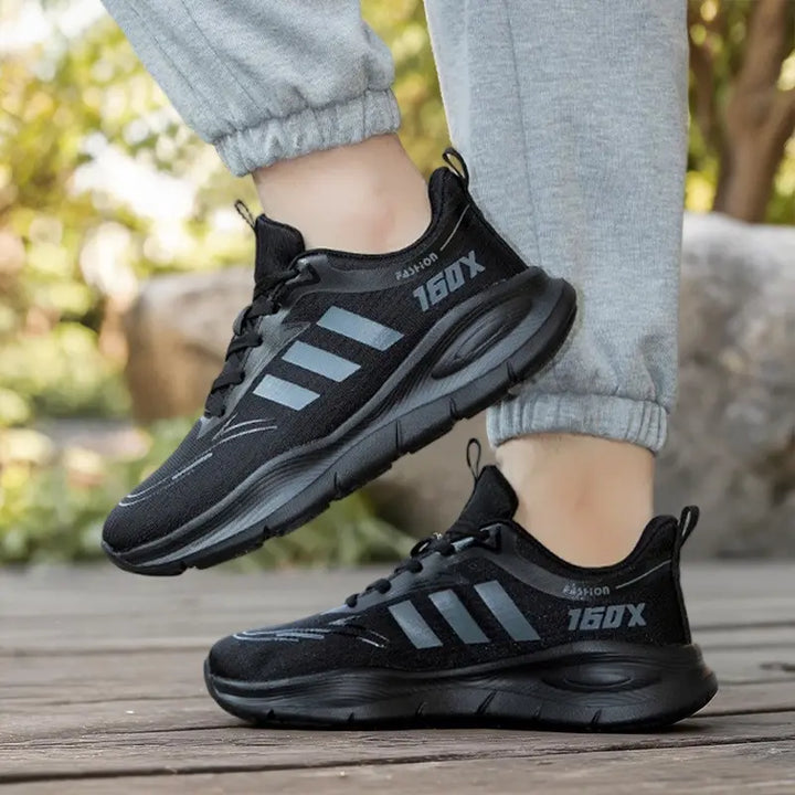 Black Trovik chunky sole sneakers with breathable mesh upper and grey stripes, shown on feet in an outdoor lifestyle setting. Features &