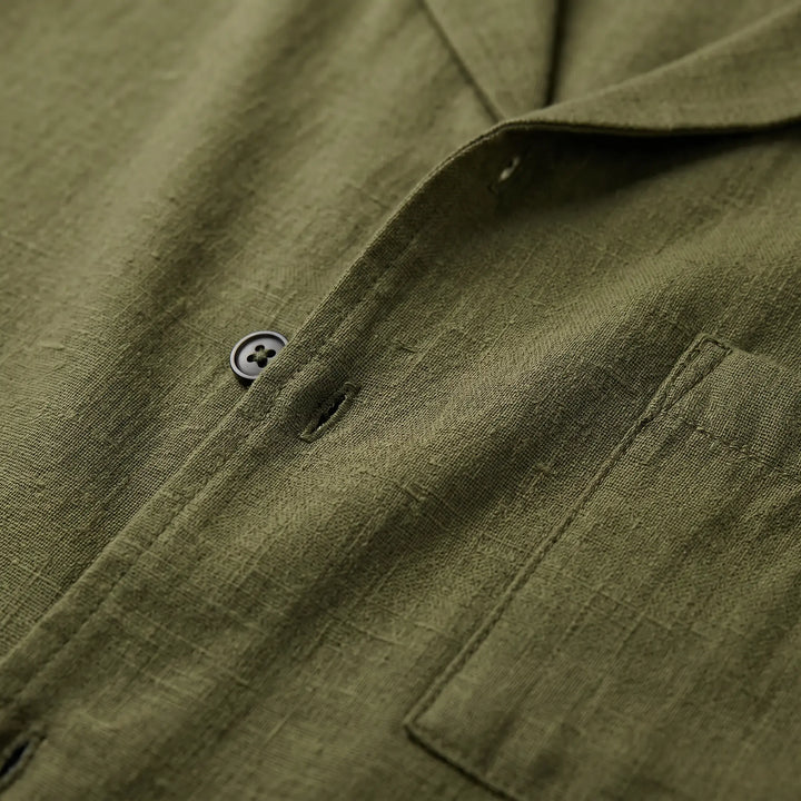 Close-up view of olive green linen-blend shirt fabric, showing matte black button, buttonhole, and chest patch pocket detail.