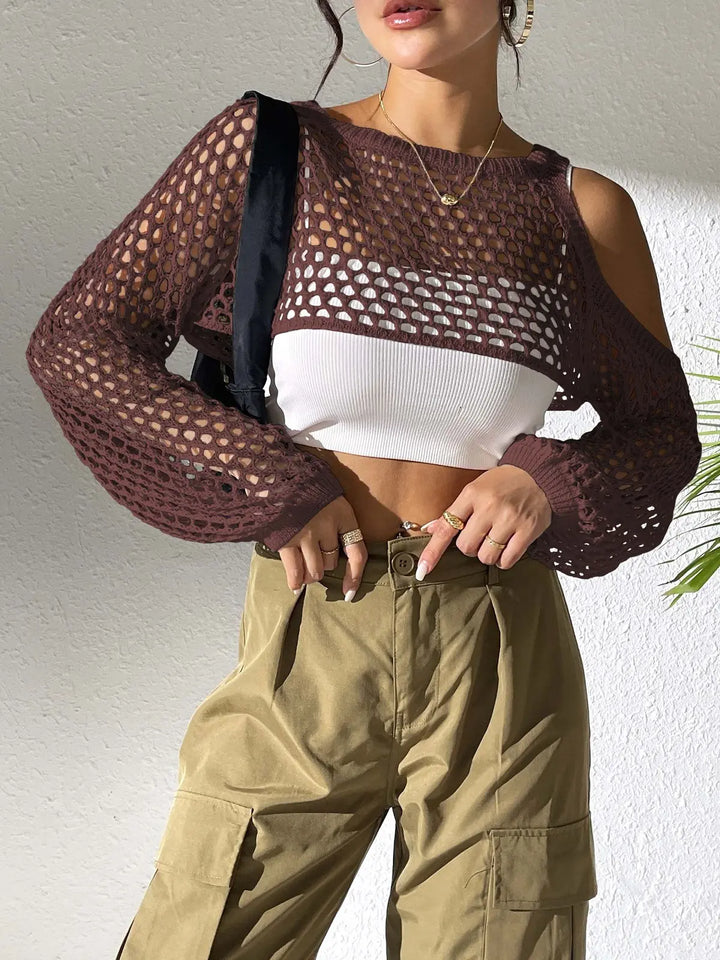 Model wearing a dark brown, open-knit crochet crop top with cold-shoulder detail and voluminous sleeves, layered over a white camisole and styled with khaki cargo pants and gold jewelry in a softly lit interior.
