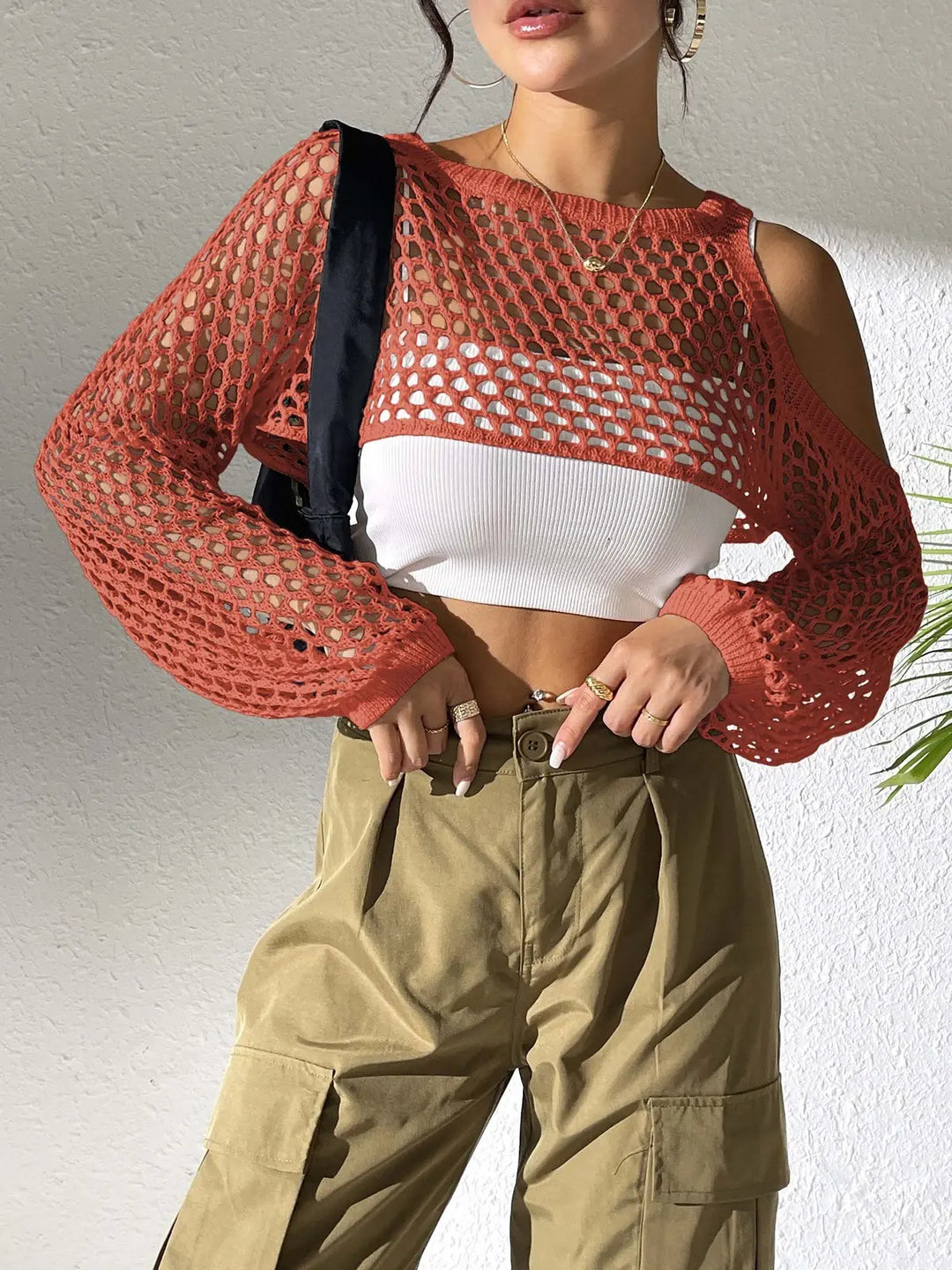 A woman wears a rust orange crochet crop top with cold-shoulder detail and voluminous sleeves over a white tank top, styled with khaki cargo pants and gold jewelry in a softly lit interior.