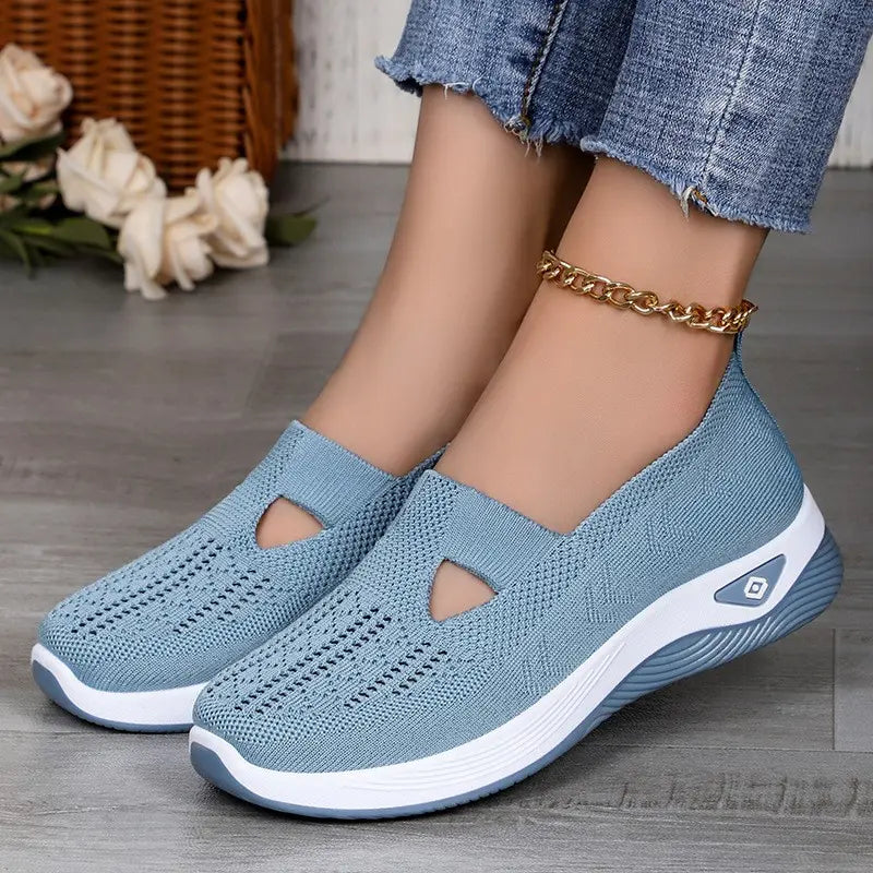Light blue Vespera breathable mesh slip-on trainers worn by a model with jeans on a wooden floor, showcasing the slip-on design and knit texture.