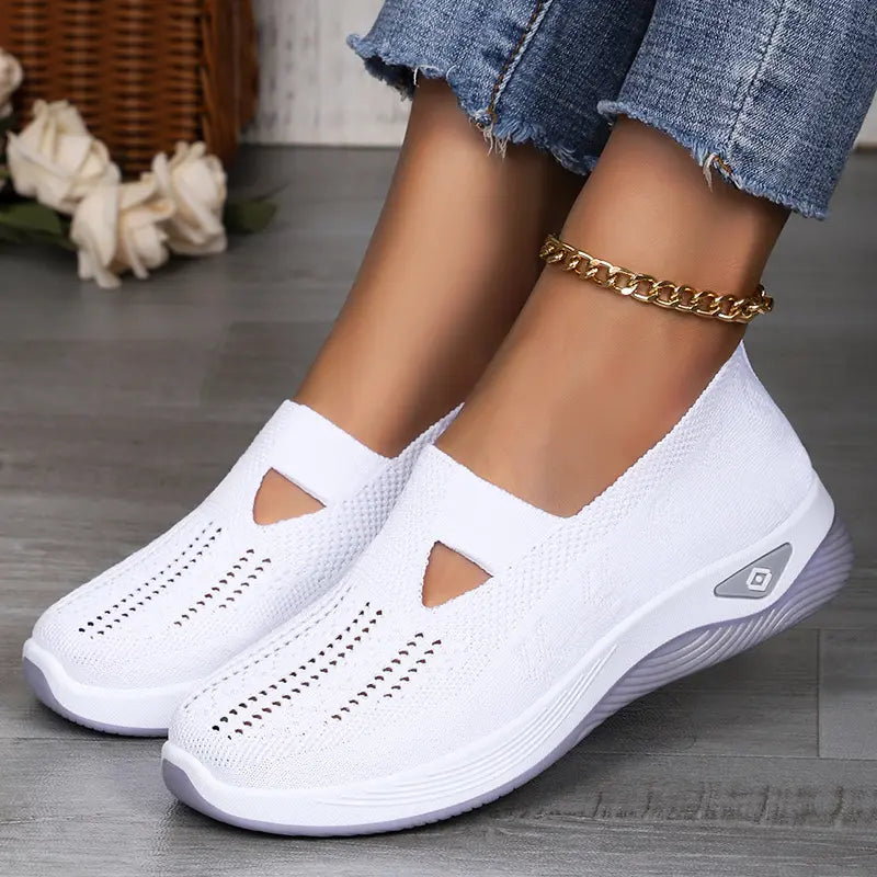 Lifestyle shot of white Vespera breathable mesh slip-on trainers worn by a person with blue jeans and a gold anklet, showcasing their comfort and casual style on a wooden floor.