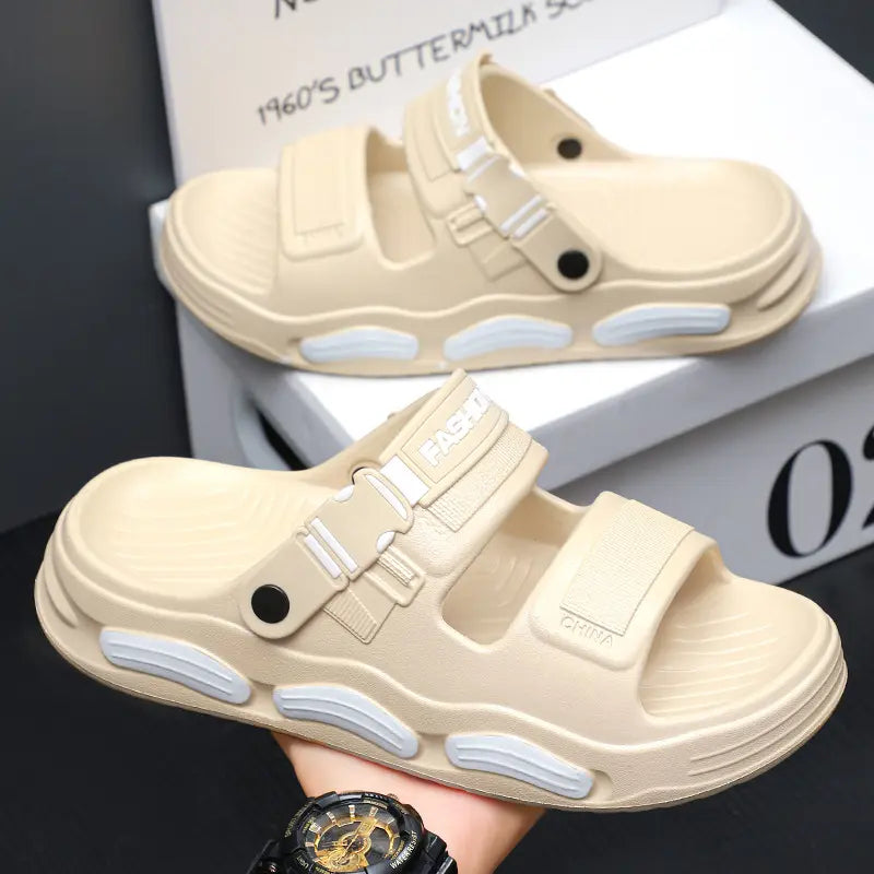 Beige men's Vespyr slide sandals shown in a three-quarter view, featuring dual adjustable straps with 'FASHION' and 'CHINA' embossing, plastic buckles, and a chunky sole with white accent pods. One sandal is held in hand against a studio backdrop.
