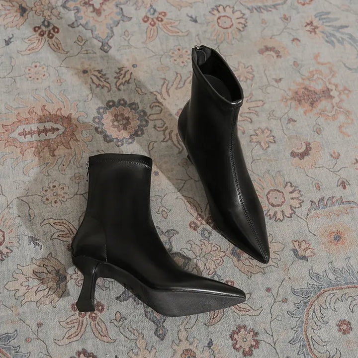 A pair of black Vesryn pointed-toe stiletto ankle boots with sculptural flared heels displayed on a patterned rug, showcasing their avant-garde design and sophisticated silhouette.