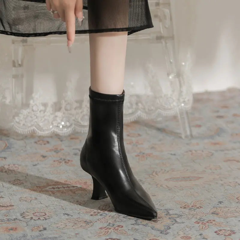 A woman's leg wearing a black pointed-toe Vesryn stiletto ankle boot with a sculptural flared heel, shown from the side.
