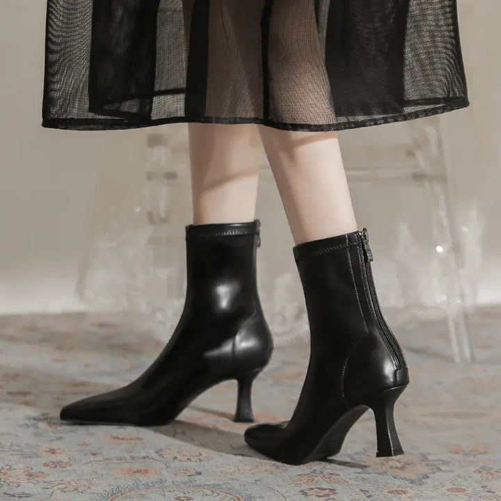 A pair of black Vesryn chic pointed toe stiletto boots with a sculptural flared heel, shown on a model from a three-quarter view with a back zipper.