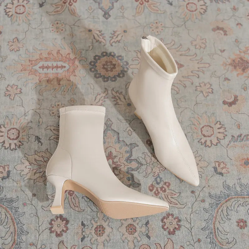 A pair of creamy off-white Vesryn pointed-toe stiletto ankle boots with sculptural flared heels, shown resting on a vintage-style patterned rug.