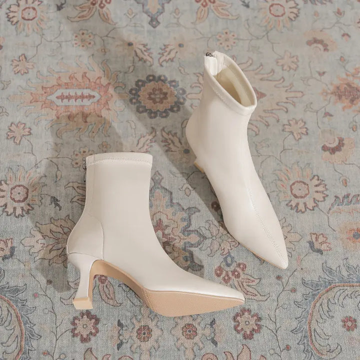 A pair of creamy off-white Vesryn pointed-toe stiletto ankle boots with sculptural flared heels, shown resting on a vintage-style patterned rug.