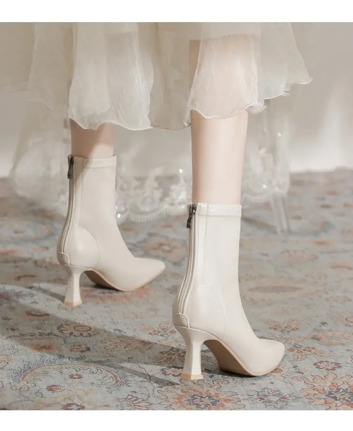 Chic creamy off-white pointed-toe stiletto ankle boots with a sculptural flared heel and back zipper, shown on a model wearing a flowing white skirt.