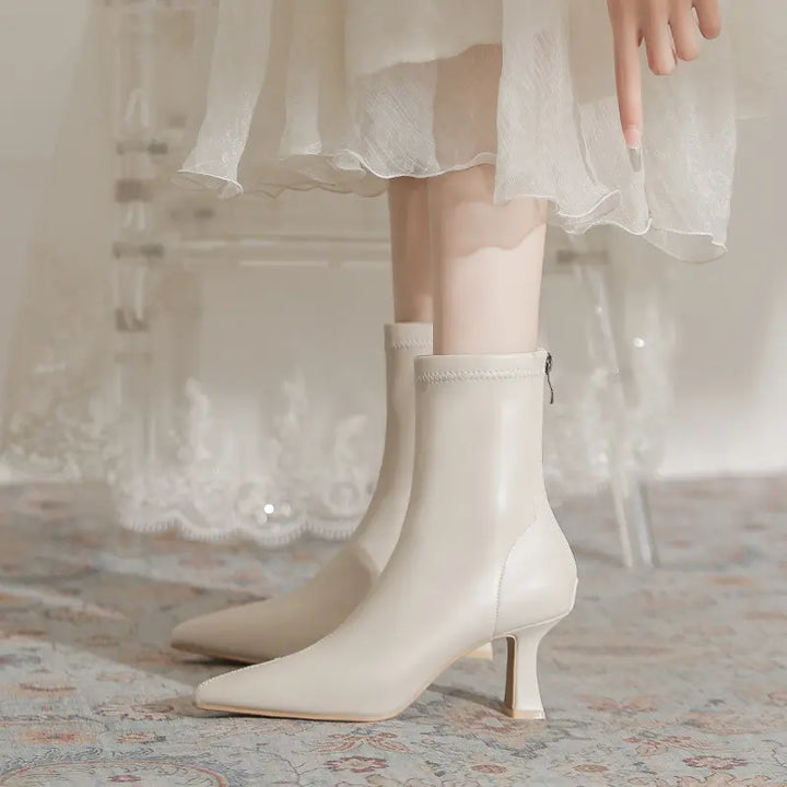 A pair of creamy off-white pointed-toe stiletto ankle boots, featuring a sculptural flared heel and back zipper, shown on legs with a delicate dress overlay.