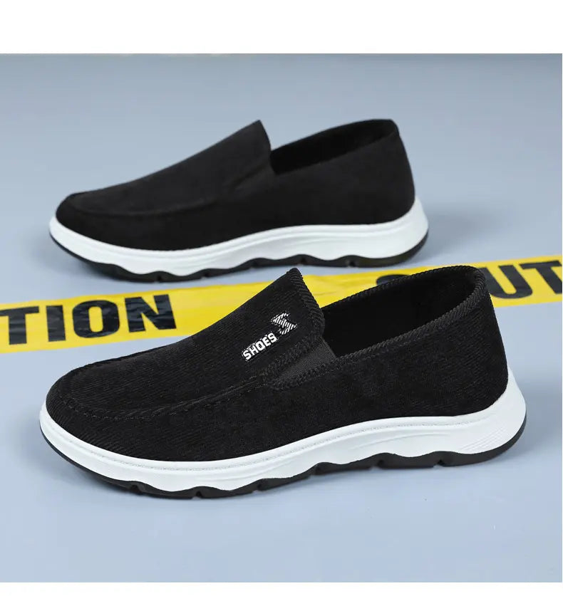 Black ribbed textile slip-on sneakers with white wavy EVA midsole and black rubber outsole, shown from a three-quarter angle on a studio background with caution tape detail.