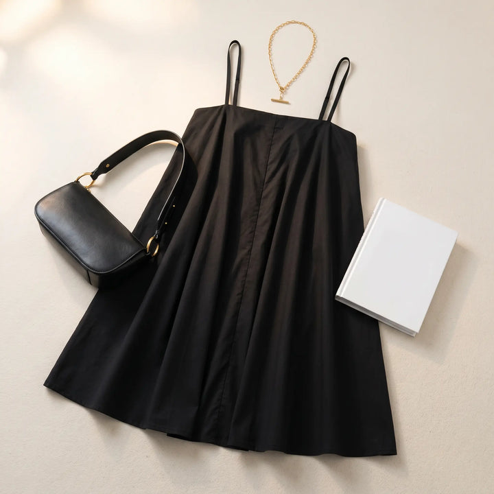 Flat lay of a black Vexana cotton sling mini dress, styled with a black shoulder bag and gold necklace, presented on a textured studio background.