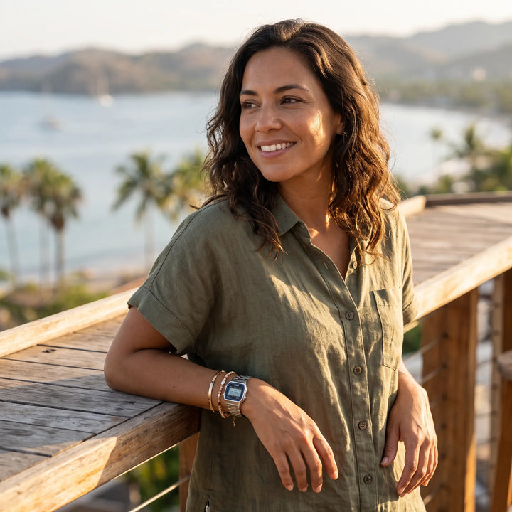A smiling woman wearing a silver Vexil retro digital stainless steel watch and bracelets, leaning on a wooden railing with a tropical ocean and palm trees in the background.