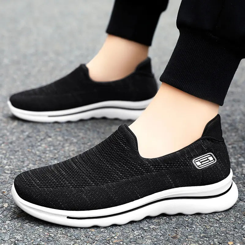 Black Veyla comfort knit slip-on trainers worn on feet, showing side view with white cushioned sole and subtle embossed side badge. These breathable walking shoes feature a modern rounded toe.