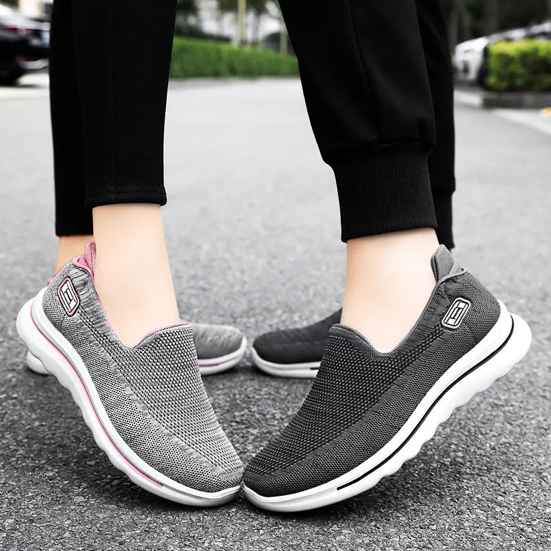 Veyla unisex slip-on trainers in grey with pink accents and dark grey, shown worn by models on a paved street. Features breathable knit upper and cushioned sole.