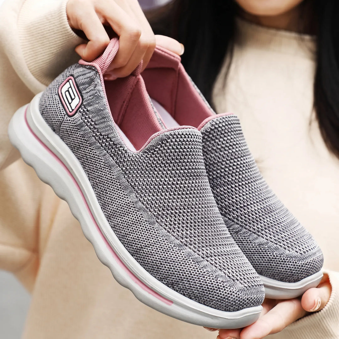 Two pairs of unisex Veyla comfort knit slip-on trainers shown in action on a street setting, one grey pair on the left and a grey pair with pink accents on the right.