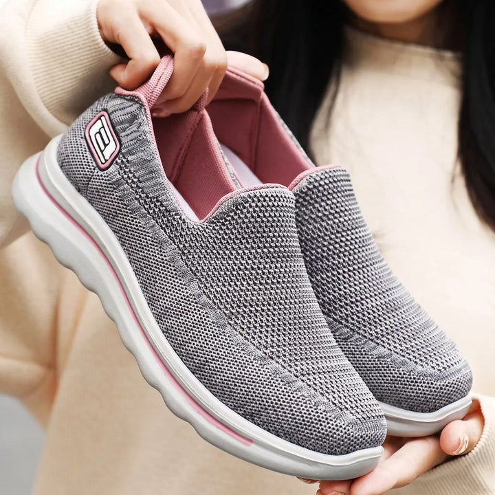 Two pairs of unisex Veyla comfort knit slip-on trainers shown in action on a street setting, one grey pair on the left and a grey pair with pink accents on the right.