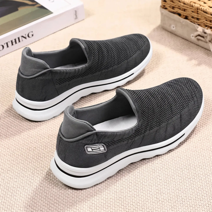 A pair of dark grey Veyla comfort knit slip-on trainers displayed on a textured surface, viewed from a three-quarter angle, showcasing their breathable mesh upper and white EVA sole with a black accent line.