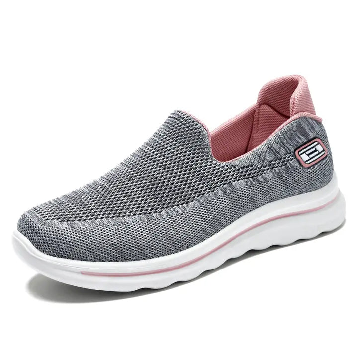 Veyla Comfort Knit Trainers in grey with pink accents, featuring a breathable slip-on design and a white EVA sole. This image shows the three-quarter side view of the sneaker on a clean white background.
