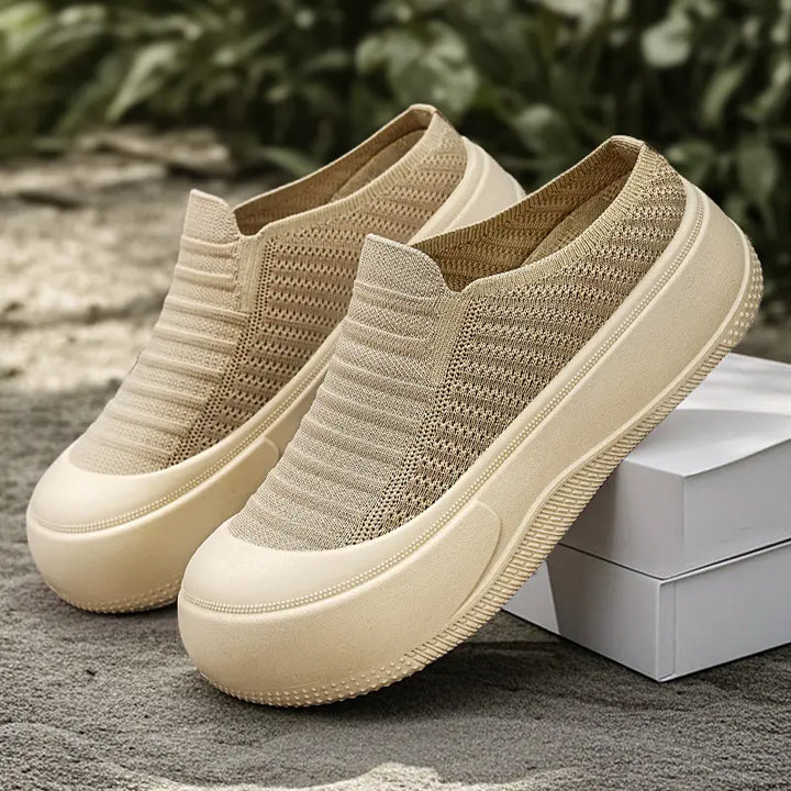 Pair of Vimera casual beige knit slip-on platform mules shown in a three-quarter view on a textured outdoor surface with greenery in the background. Features a round toe and thick, light-colored sole.