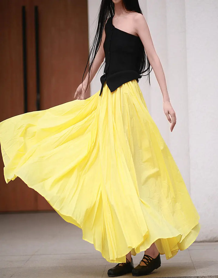 A woman models a flowing, voluminous yellow maxi skirt with a black one-shoulder top, showcasing its dramatic sweep and bohemian style.