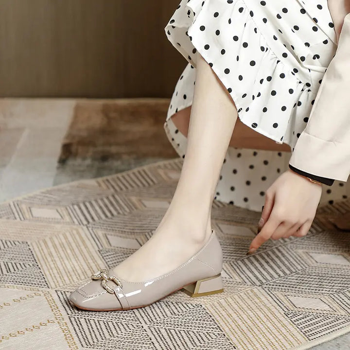 A model wears elegant taupe patent leather square-toe pumps with a decorative gold buckle embellishment, paired with a polka dot dress.