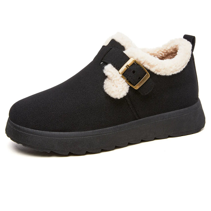 Vylos black ankle boot featuring a plush cream faux fur lining, decorative brass buckle strap with pom-poms, and a durable lugged sole, shown from a side view.