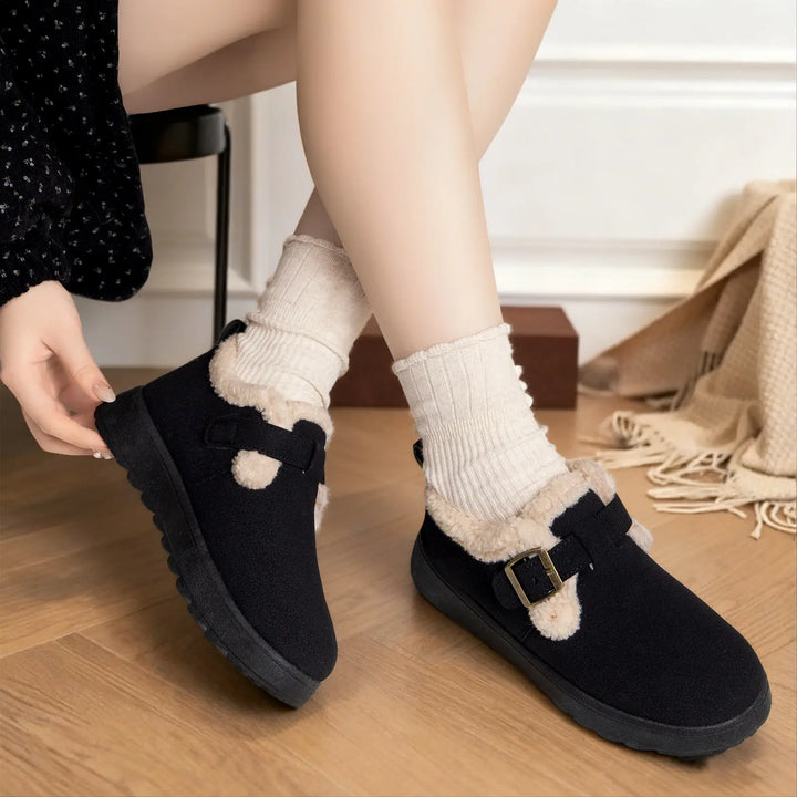Black Vylos ankle boots featuring plush faux fur lining and an adjustable buckle strap with pom-pom accents, worn by a model with cream ribbed socks on a wooden floor.