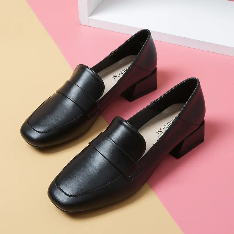 Black Vylos square-toe loafers with a decorative vamp strap and block heel, shown from a front-three-quarter angle on a pastel pink and yellow background.