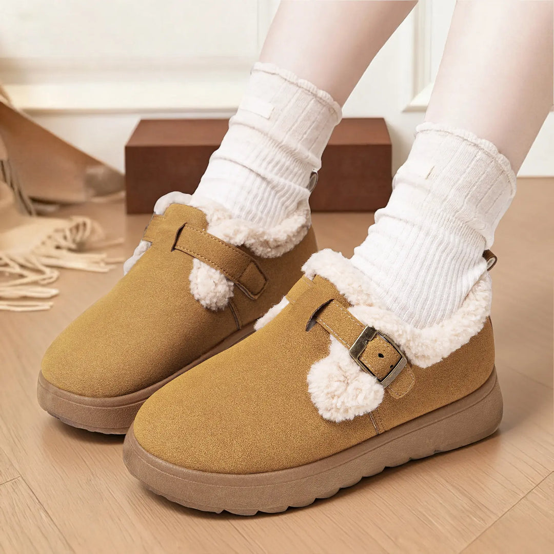 Camel suede-like women's winter boots with plush faux fur lining, decorative buckle straps, and pom-poms, shown on a model with white ribbed socks on a wooden floor.