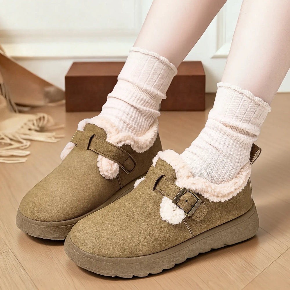 Close-up of Vylos women's khaki winter boots with plush faux fur lining and decorative buckle strap, worn by a model with cream ribbed socks on a wooden floor.