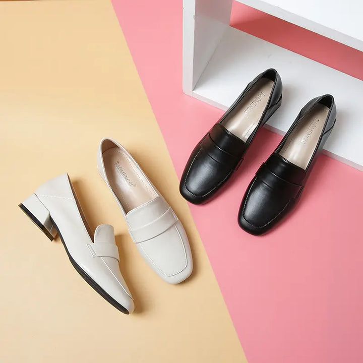 Studio arrangement of black and off-white Vylos square-toe loafers, showcasing their slip-on design, decorative vamp strap, and block heels on a split yellow and pink background.