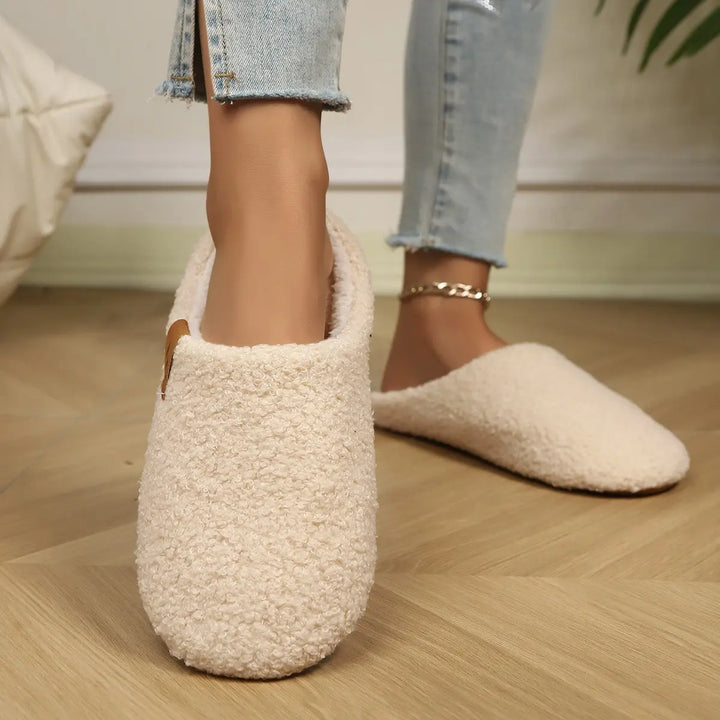 A pair of cream beige Vyrra plush indoor clog slippers shown worn on feet with light wash jeans and a gold anklet, demonstrating cozy comfort and warm home wear on a wooden floor.