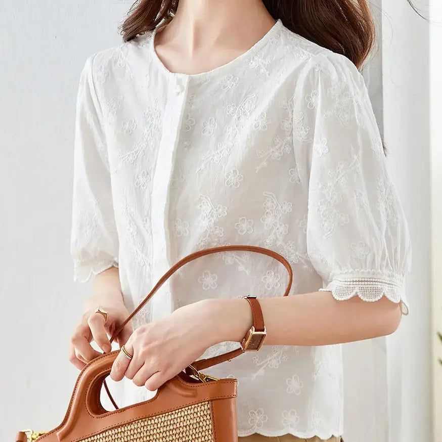 A woman wearing a white embroidered cotton blouse with scalloped lace trim on puff sleeves and hem, paired with tan trousers and a woven tote bag.