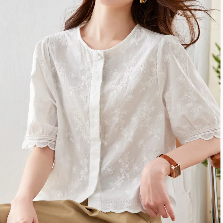 A woman wears a chic white cotton blouse featuring all-over floral embroidery, scalloped lace trim on short puff sleeves, and a round button-front neckline, styled with tan trousers.