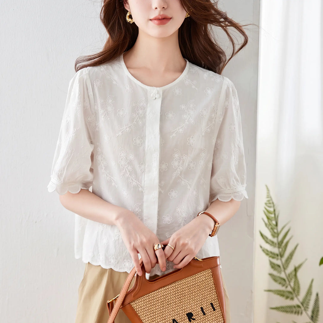 Front view of a white embroidered cotton blouse with a round neckline, puff sleeves, and scalloped lace trim. This chic summer top features delicate floral embroidery all over and a button-front closure.