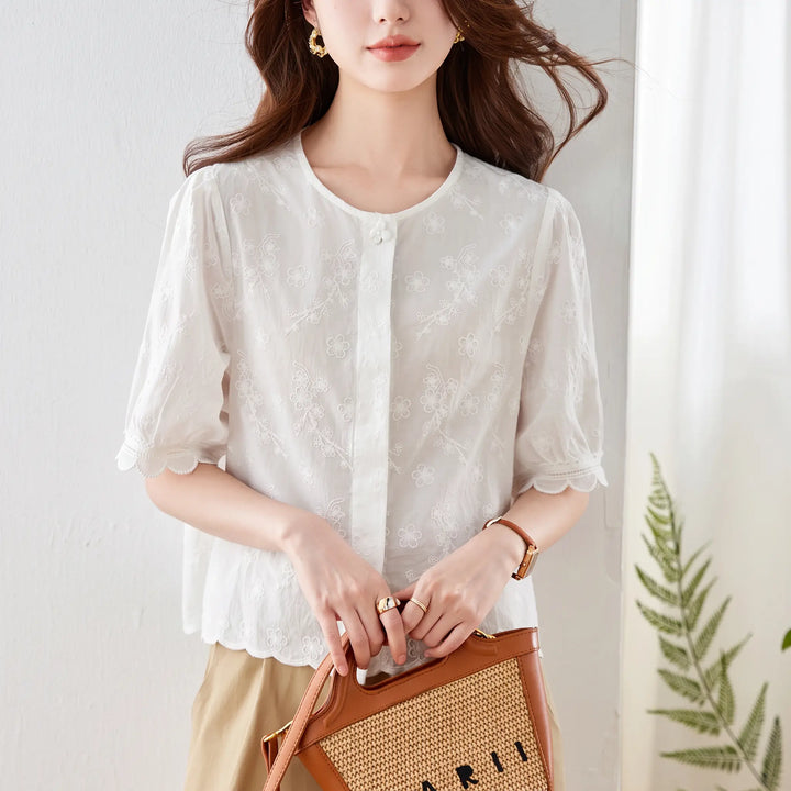 Front view of a white embroidered cotton blouse with a round neckline, puff sleeves, and scalloped lace trim. This chic summer top features delicate floral embroidery all over and a button-front closure.