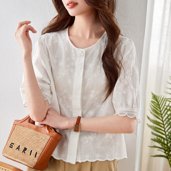 Chic white embroidered cotton blouse with scalloped lace trim on puff sleeves and hem, worn by a model holding a straw bag and watch, styled for summer.