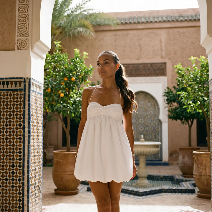 A woman models a white halterneck mini dress with a ruched bodice and voluminous bubble hem, standing in a sunlit courtyard with traditional Moroccan-style tiles and lush greenery.