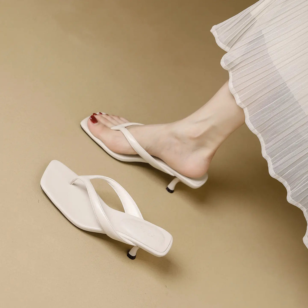 A pair of white heeled flip-flops with a square toe and stiletto heel, shown on a foot with a white pleated skirt detail.