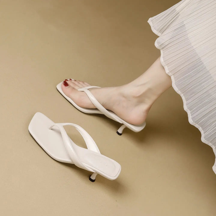 A pair of white heeled flip-flops with a square toe and stiletto heel, shown on a foot with a white pleated skirt detail.