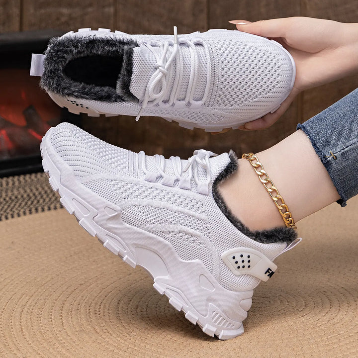 A pair of white Kryptik knit ankle boots with plush faux fur lining and a chunky platform sole, shown on a foot with a gold anklet and another boot held by hand, styled for winter comfort.