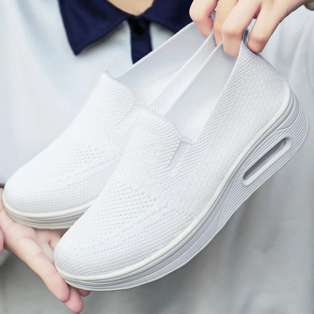 Pair of white Xylos breathable knit slip-on sneakers with a visible air cushion sole, held by hands against a blurred background.