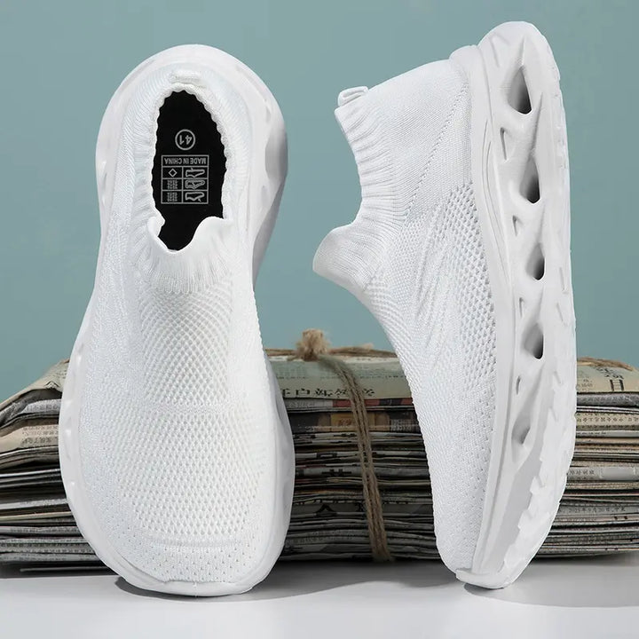 Pair of white slip-on knit trainers with a sculpted wave-patterned sole, shown from a three-quarter angle with the left shoe forward.