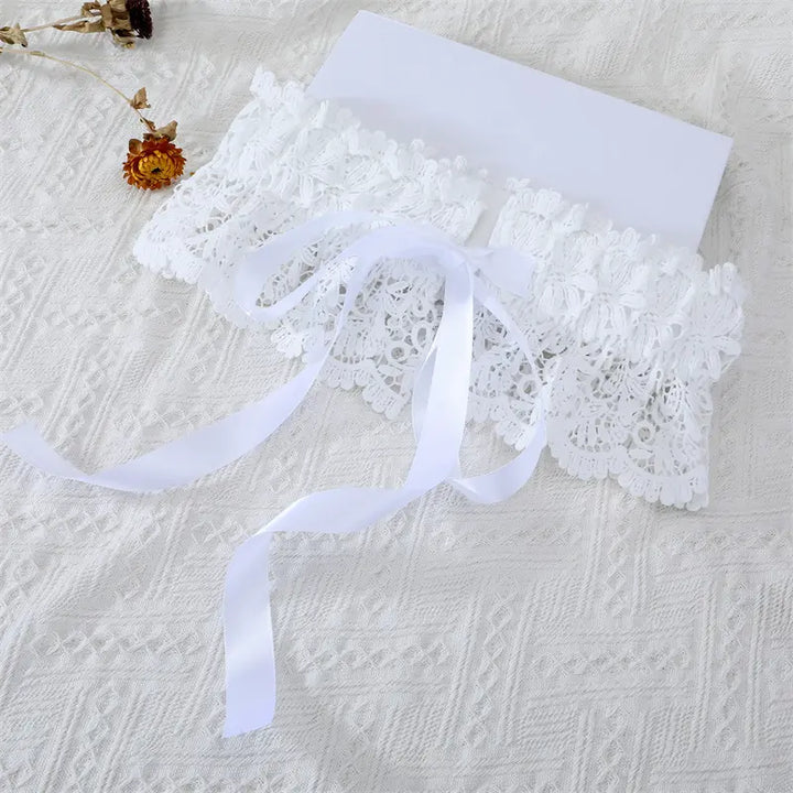 White floral lace cummerbund belt with wide satin ribbon ties, laid flat on a textured white surface with decorative dried flowers.