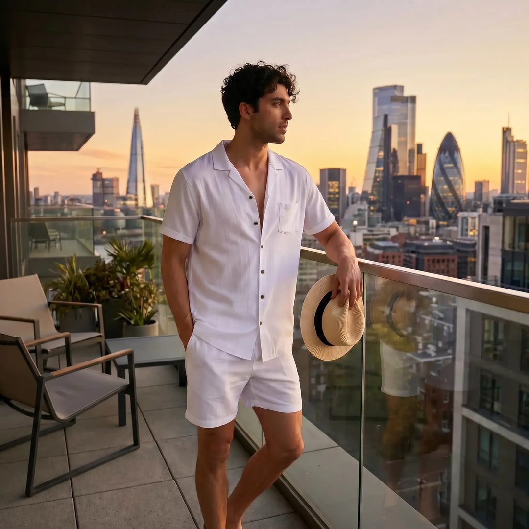 Man wearing a white linen-blend short-sleeved camp collar shirt and matching shorts on a balcony overlooking a city skyline at sunset.