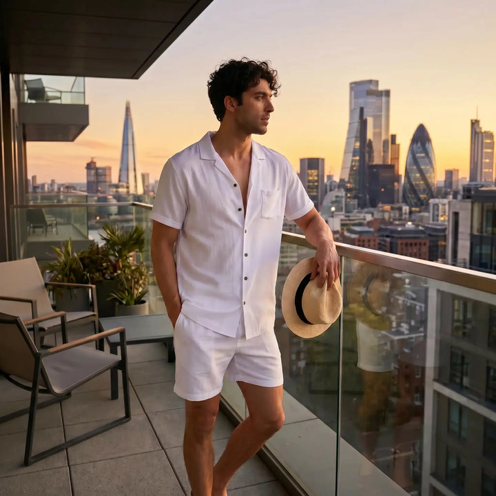 Man wearing a white linen-blend short-sleeved camp collar shirt and matching shorts on a balcony overlooking a city skyline at sunset.